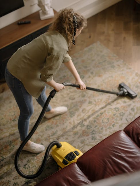 A person is kneeling on a patterned carpet, using a yellow and black vacuum cleaner labeled 'North Sheen Carpet Cleaners' to perform deep cleaning and surface sanitisation. Their hands are placing a w