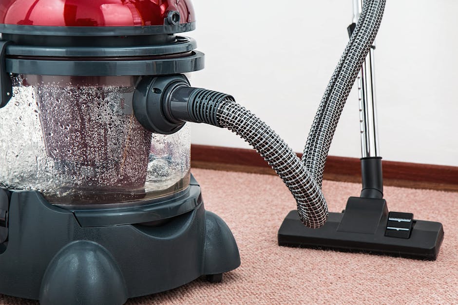 A professional cleaner dressed in a white protective suit, gloves, face mask, and shoe covers is performing surface cleaning on a beige, plush carpet using a vacuum cleaner with a long hose attachment